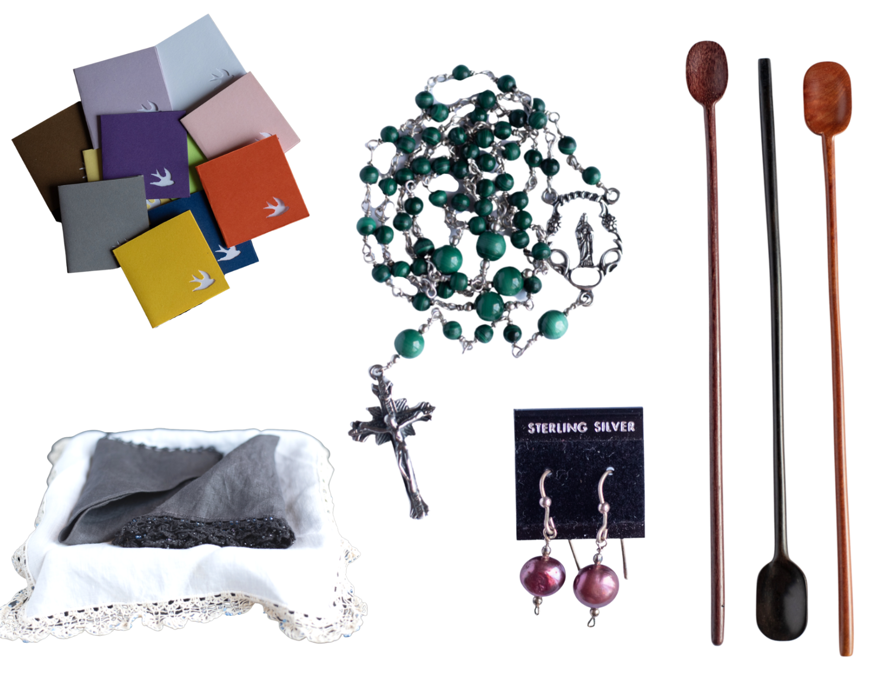 The photo shows several two inch by two inch linen lined note cards with a flying dove punched out, a coiled malachite and sterling rosary, black and white linen hand laced handkerchiefs. a pair of sterling and burgundy pearl earrings on an a black earring card and three polished long wooden teaspoons in purple heart, ebony and pink ivory woods. arranged on a gray gingham background.