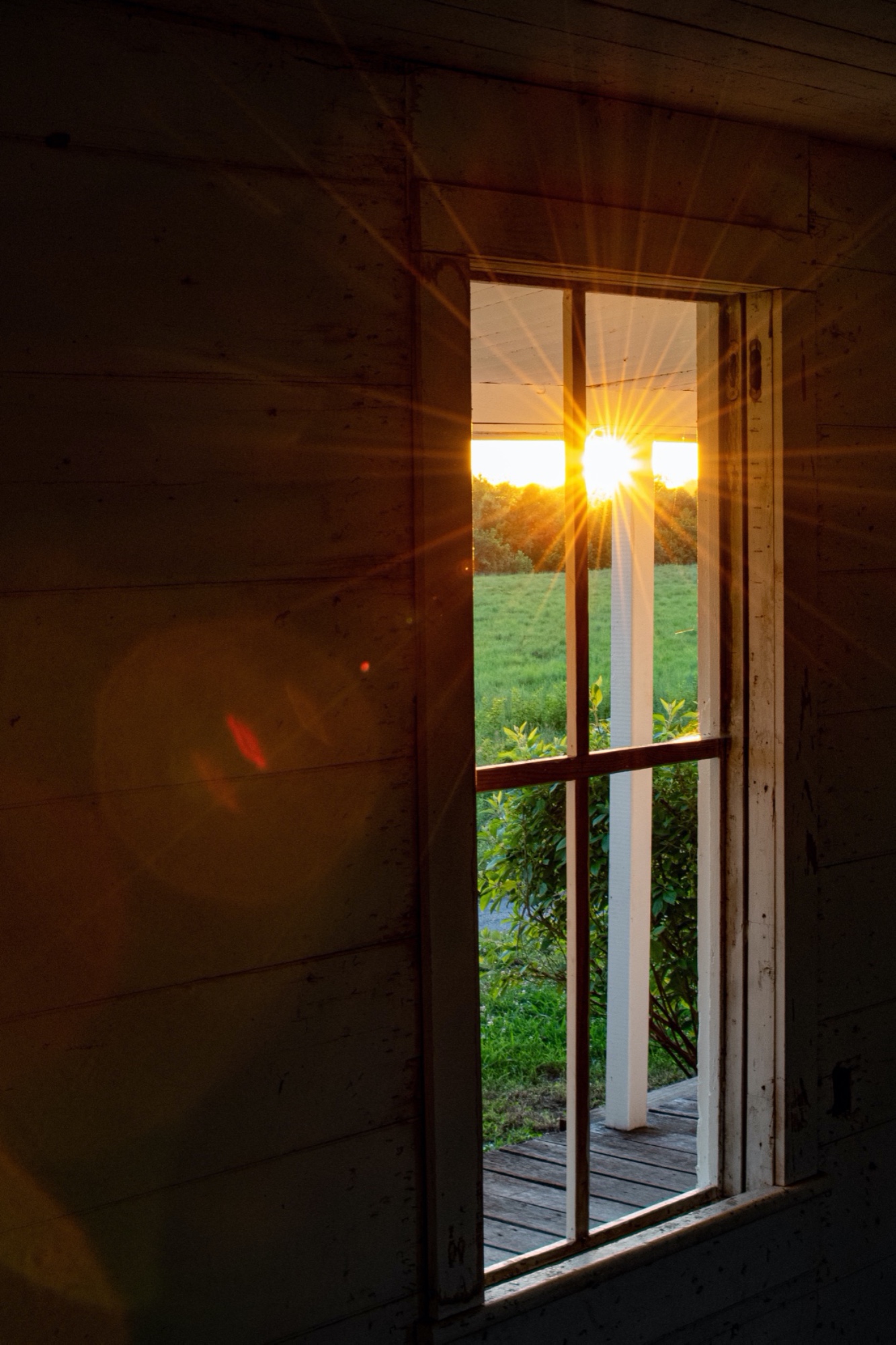 The Photo shows a window, through which a setting sun can be seen, over low crops in a field.